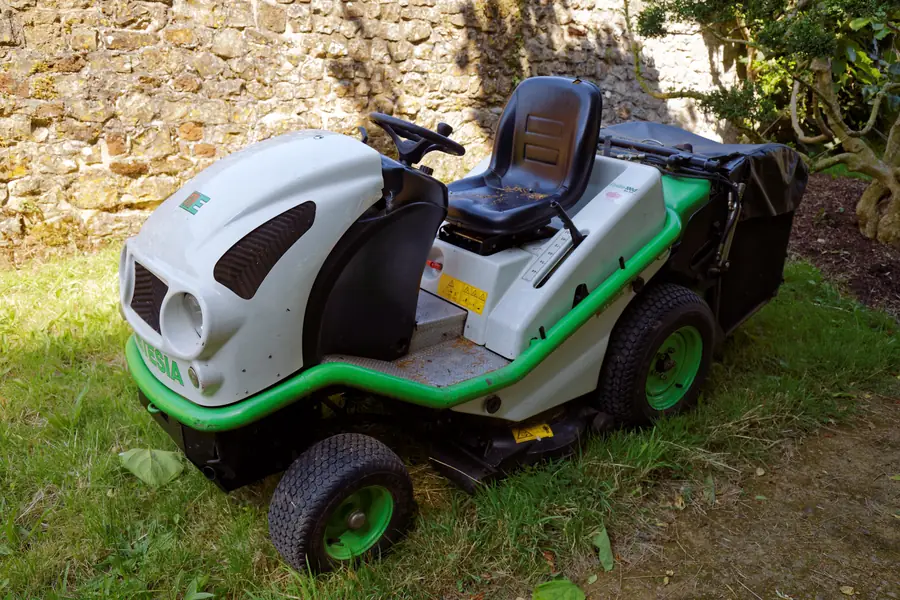 Green and white ride-on lawn mower parked on grass beside a stone wall at Parham Park, West Sussex