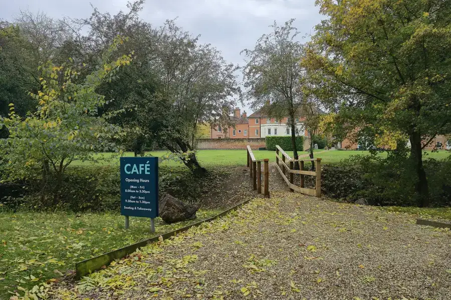 Path and lawn edged by mature trees, with a café sign, at Thremhall Park, Essex